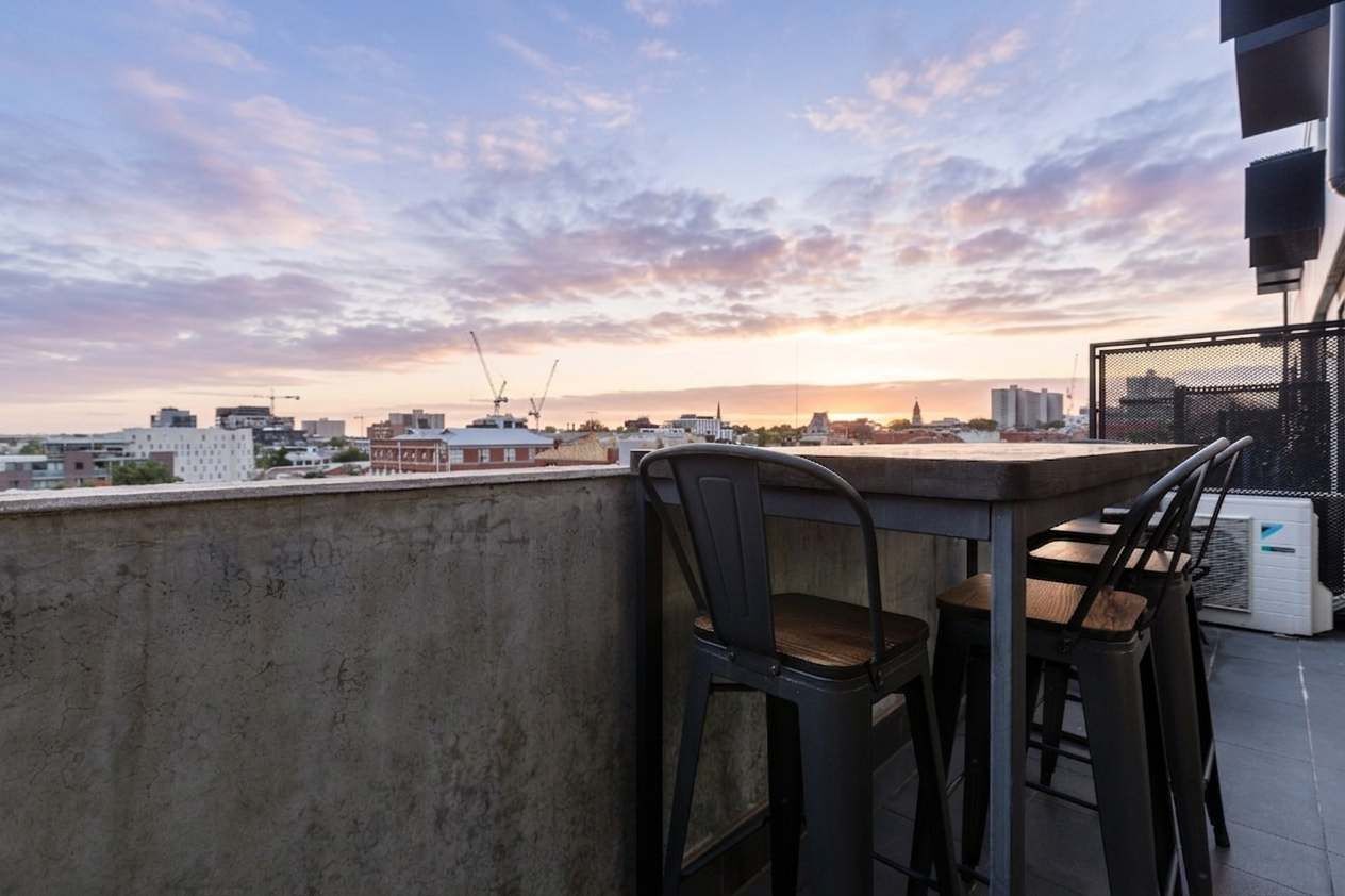 Terraza en la azotea con sillas y una mesa, con vistas a un paisaje urbano al atardecer.