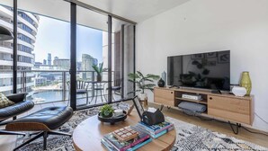 Una sala de estar moderna con un televisor grande, un sofá, una mesa de centro con libros y vistas a la ciudad a través de amplias ventanas.