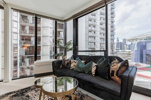 A modern living room with a black sofa, a glass coffee table, and a large window with a city view. - Sleek glamour steps from Marvel Stadium and marina (Docklands)