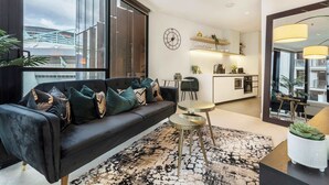 A modern living room with a sofa, a coffee table, and a rug. There is a kitchen area in the background with a dining table and chairs.