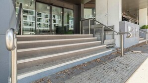 A modern building entrance with a staircase leading to a glass door, metal handrails, and a tactile paving path.