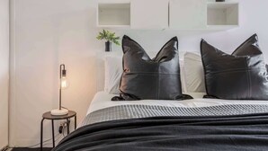 A neatly made bed with a quilted blanket, two black pillows, a bedside lamp, and a small potted plant on a shelf above the bed.