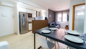 A modern kitchen with a refrigerator, microwave, and wooden panel divider. There is a dining table set with plates, glasses, and cutlery. A gray sofa is in the background, and a window with blinds is visible. - Up to 5 Guests, Garage, Elevator, Swimming Pool, Jr Catito (Brasília)