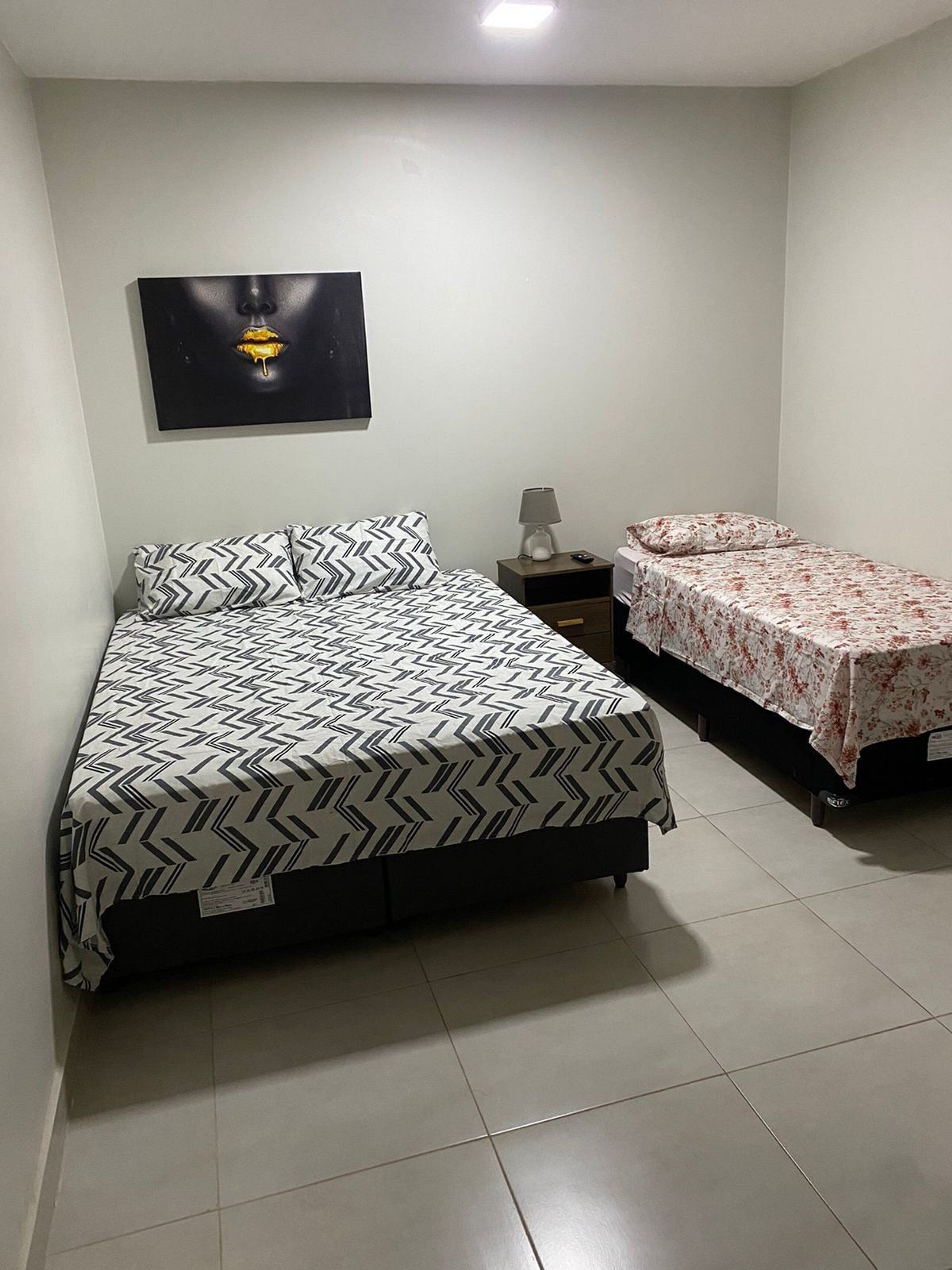 Une chambre à coucher avec un grand lit, un plus petit lit, une table de chevet, une lampe et un tableau encadré au mur.