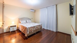 A bedroom with a wooden floor, a bed with floral bedding, a nightstand with a lamp, and a window with white curtains.