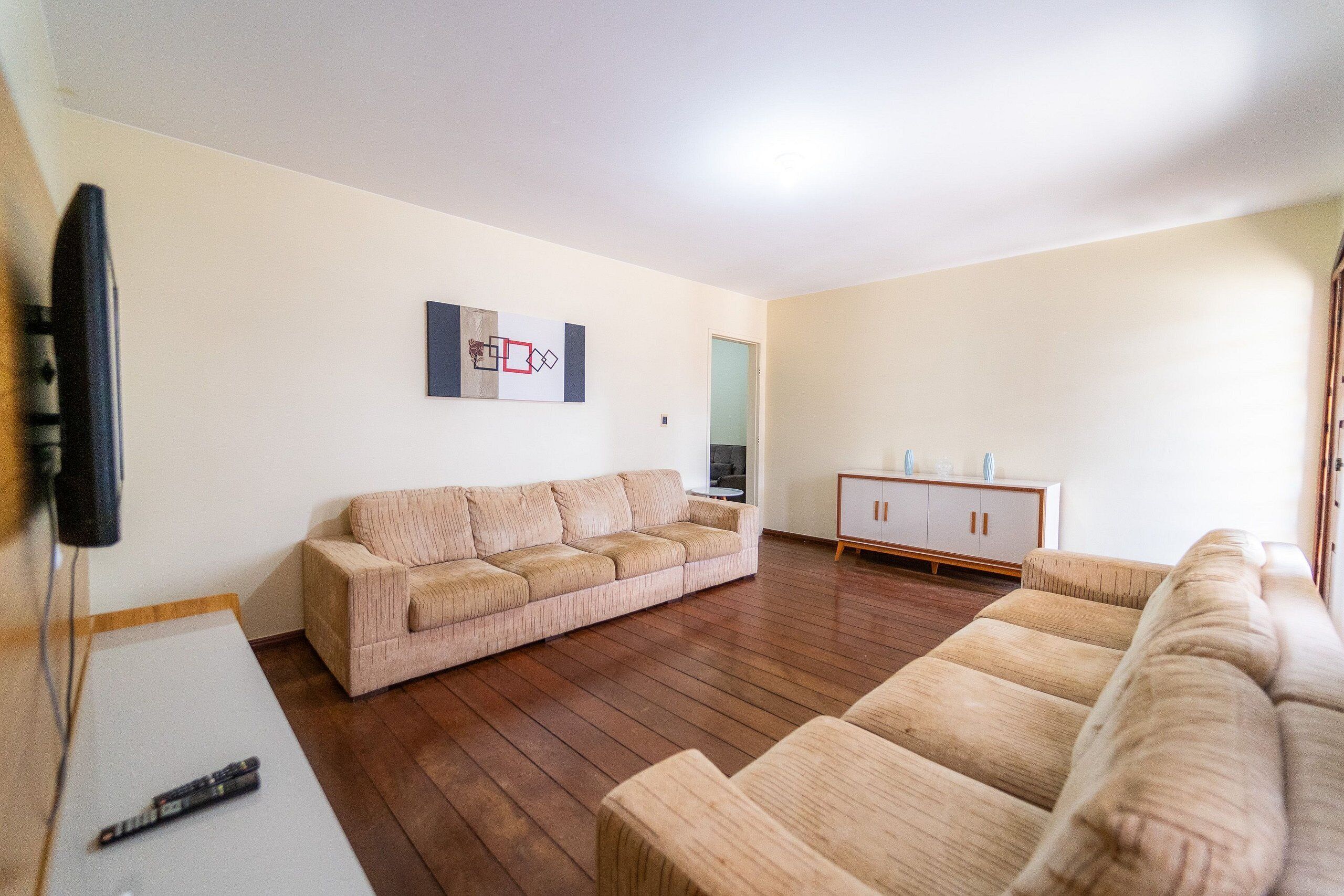 A living room with a beige sofa, a wooden floor, a cabinet, and a flat-screen TV mounted on the wall.