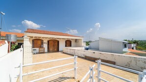 A rooftop terrace with a tiled floor, a white railing, and a building with a red-tiled roof and wooden doors.