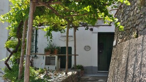 Ein schmaler Gang mit einer Holzw pergola, die dicht mit Grün bewachsen ist, einer Steinmauer und einem weißen Gebäude mit grüner Tür und Fenstern.