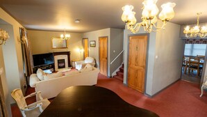 Un salon spacieux avec une grande table à manger en bois foncé, un canapé blanc et une cheminée. On y trouve deux lustres et un escalier menant à une autre pièce.