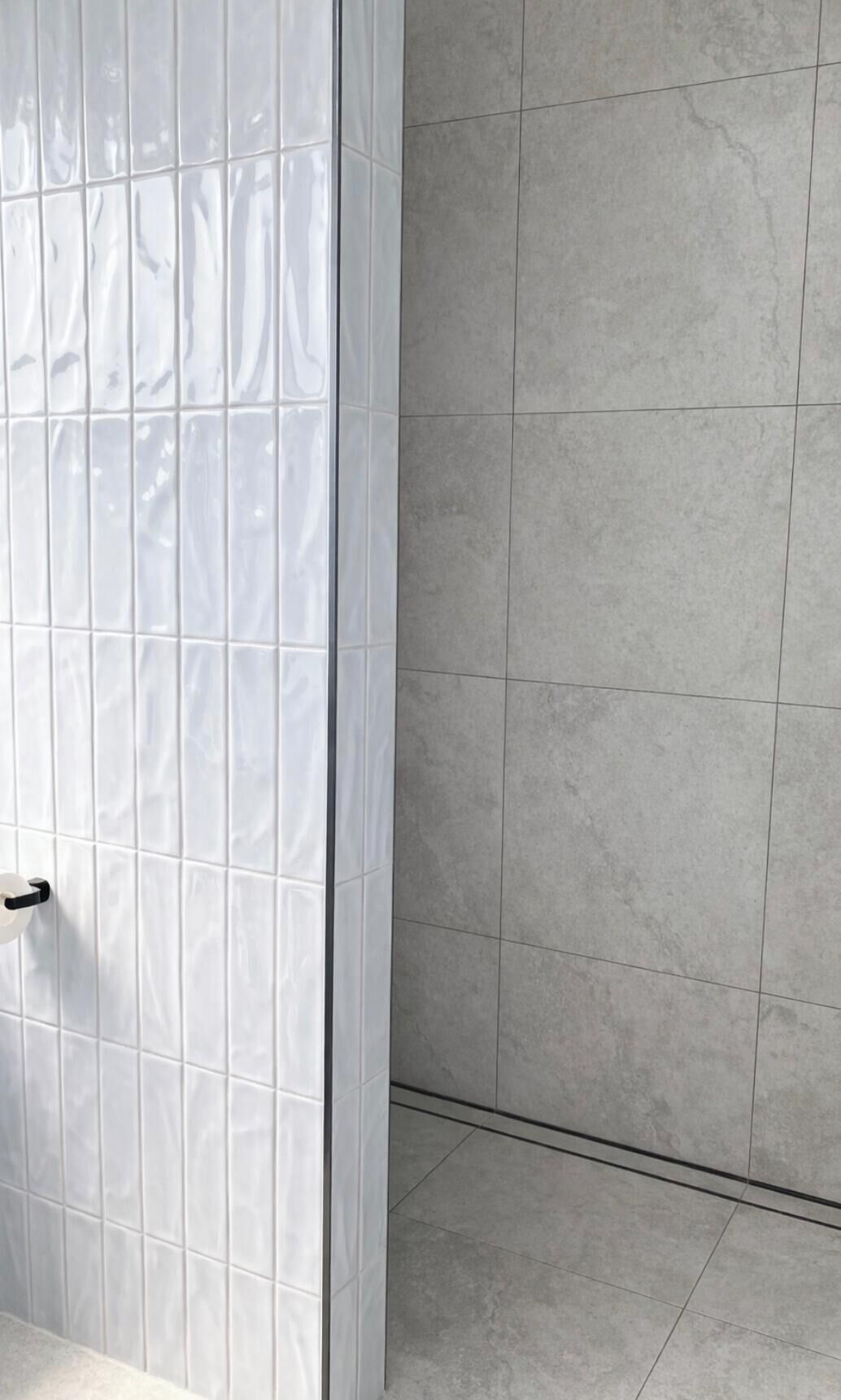 A bathroom with white tiled walls and floor, a white door with a black handle, and a shower area with grey tiles.