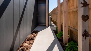 A narrow pathway leading to a dark entrance, flanked by wooden fences and metal railings, with decorative metal plaques and a bird sculpture. - Cosy ski retreat on The Remarkables Ski Area. 5 minutes to Jacks Point Golf Club (Jacks Point)