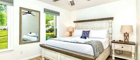 A bedroom with a large bed, two bedside tables, a ceiling fan, and a window with a view of greenery.