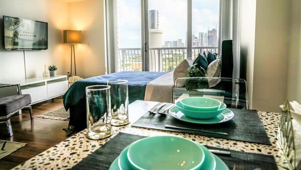 A modern hotel room with a bed, a dining table set with green bowls, a TV on the wall, and a balcony with a city view. - Amazing Studio Downtown MIA (Miami)