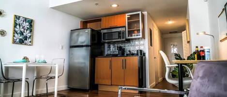 A compact kitchen with a refrigerator, microwave, and wooden cabinets. There is a dining table with chairs and a pendant light above it.