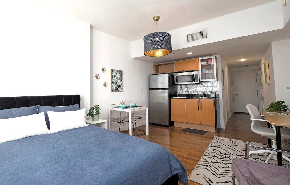 Un dormitorio moderno con una cama grande, una mesita pequeña y una kitchenette al fondo.