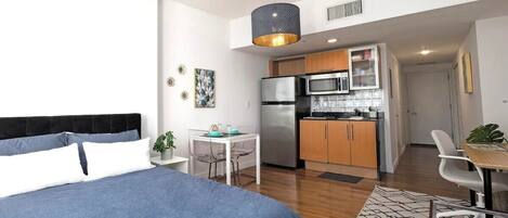 Un dormitorio moderno con una cama grande, una mesita pequeña y una kitchenette al fondo.
