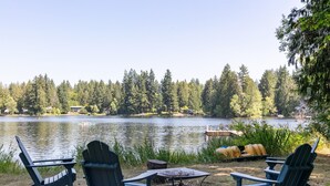 Blick auf einen See mit drei Adirondack-Stühlen, einer kleinen Feuerstelle und einem Steg, der ins Wasser ragt.