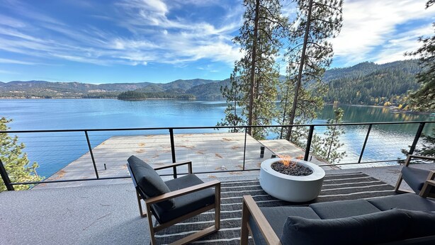 A terrace with a fire pit, two black lounge chairs, and a view of a lake and mountains.