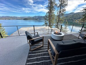 A terrace with a fire pit, two black lounge chairs, and a view of a lake and mountains. - Luxury Lake View with Sauna (Hayden)