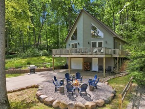 Une maison à deux étages dotée d’une terrasse, d’un espace extérieur aménagé avec des chaises et un foyer, le tout entouré d’une zone recouverte de gravier et de gros rochers.