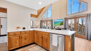 Cocina moderna con armarios de madera, un refrigerador de acero inoxidable y ventanas amplias con vistas a árboles y un paisaje.