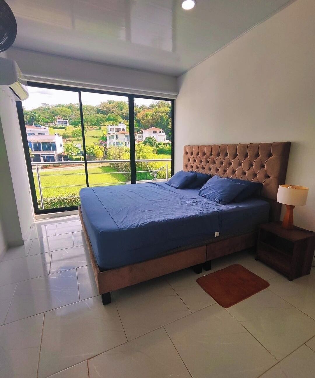 A bedroom with a large bed, a nightstand, a lamp, and a view of greenery and buildings through a large window.