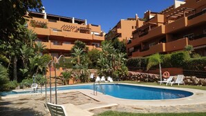 A swimming pool with lounge chairs, surrounded by palm trees and a multi-story building with balconies.