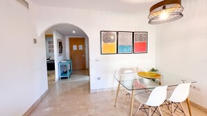 A modern dining area with a glass table and chairs, a hanging light fixture, and a wall with abstract art.