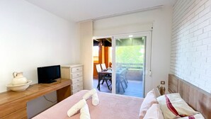 A hotel room with a bed, a TV, a desk with a bowl and a remote, and a sliding glass door to an outdoor area with a table and chairs.