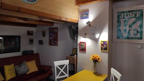 A cozy living room with a wooden ceiling, a yellow table, white chairs, a red couch with yellow cushions, a wall clock, and various framed pictures and decorations.