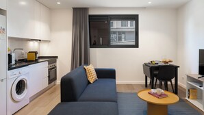 Une cuisine moderne équipée d’un lave-linge intégré, un coin repas avec une table et des chaises, et une grande fenêtre donnant sur des immeubles.