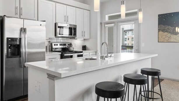 A modern kitchen with white cabinetry, stainless steel appliances, a large island with bar stools, pendant lighting, and a view through a window. - LUX 2B2B Apt Near Heart of Austin w/Pool & Parking (Austin)