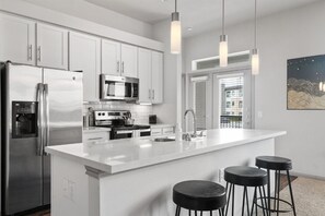 Una cocina moderna con armarios blancos, electrodomésticos de acero inoxidable, una isla grande con taburetes altos, iluminación colgante y vistas a través de una ventana.