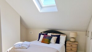 Une chambre avec un puits de lumière, un lit recouvert d’une couvre-lit beige, un coussin coloré, une lampe de chevet et une petite table de nuit en bois.