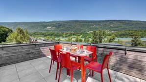 A terrace with red chairs and a table set for dining, overlooking a scenic landscape with hills and a lake.