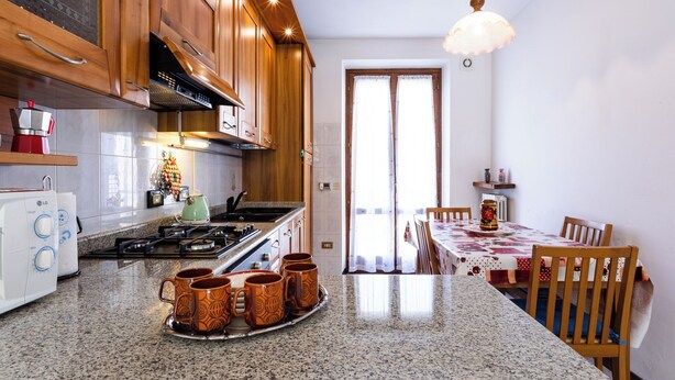 Een keuken met granieten aanrechtbladen, houten keukenkastjes, een gasfornuis en een eethoek met een tafel en stoelen.