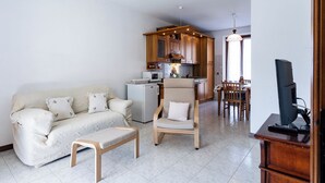 Een woonkamer met een bank, een stoel en een bijzettafeltje. Achtergrond is een keuken met houten kastjes en een eethoek met een tafel en stoelen.