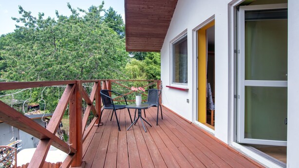 Una terraza de madera con dos sillas, una mesa, barandilla de madera y una puerta con marco amarillo.