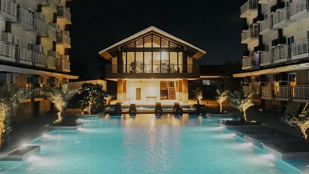A modern hotel with a swimming pool, multiple balconies, and a well-lit entrance. - Experience affordable luxury in our cozy & homey staycation layover pad.  (Lapu-Lapu City)