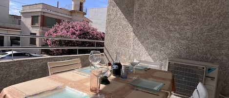 Un espace de restauration en plein air avec une table dressée pour deux personnes, des chaises blanches et une vue sur un bâtiment doté d’une tour et orné de fleurs roses.
