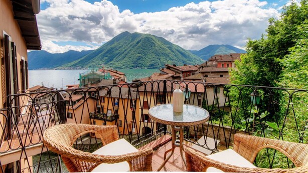 Ein Balkon mit Korbstühlen und einem kleinen Tisch, der auf eine Berglandschaft und ein Gewässer blickt.