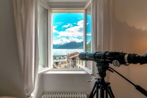 Een telescoop op een statief voor een raam, met uitzicht op gebouwen en bergen.