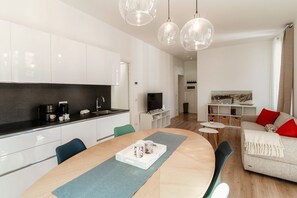 A modern kitchen with white cabinets, a dining table with chairs, and a living area with a sofa and TV.