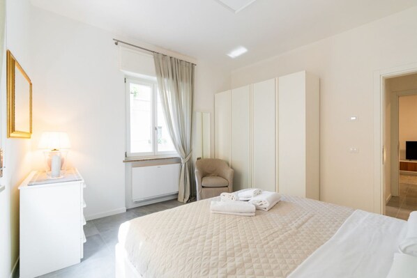 A hotel room with a bed, a chair, a desk with a lamp, a wardrobe, and a window with curtains.