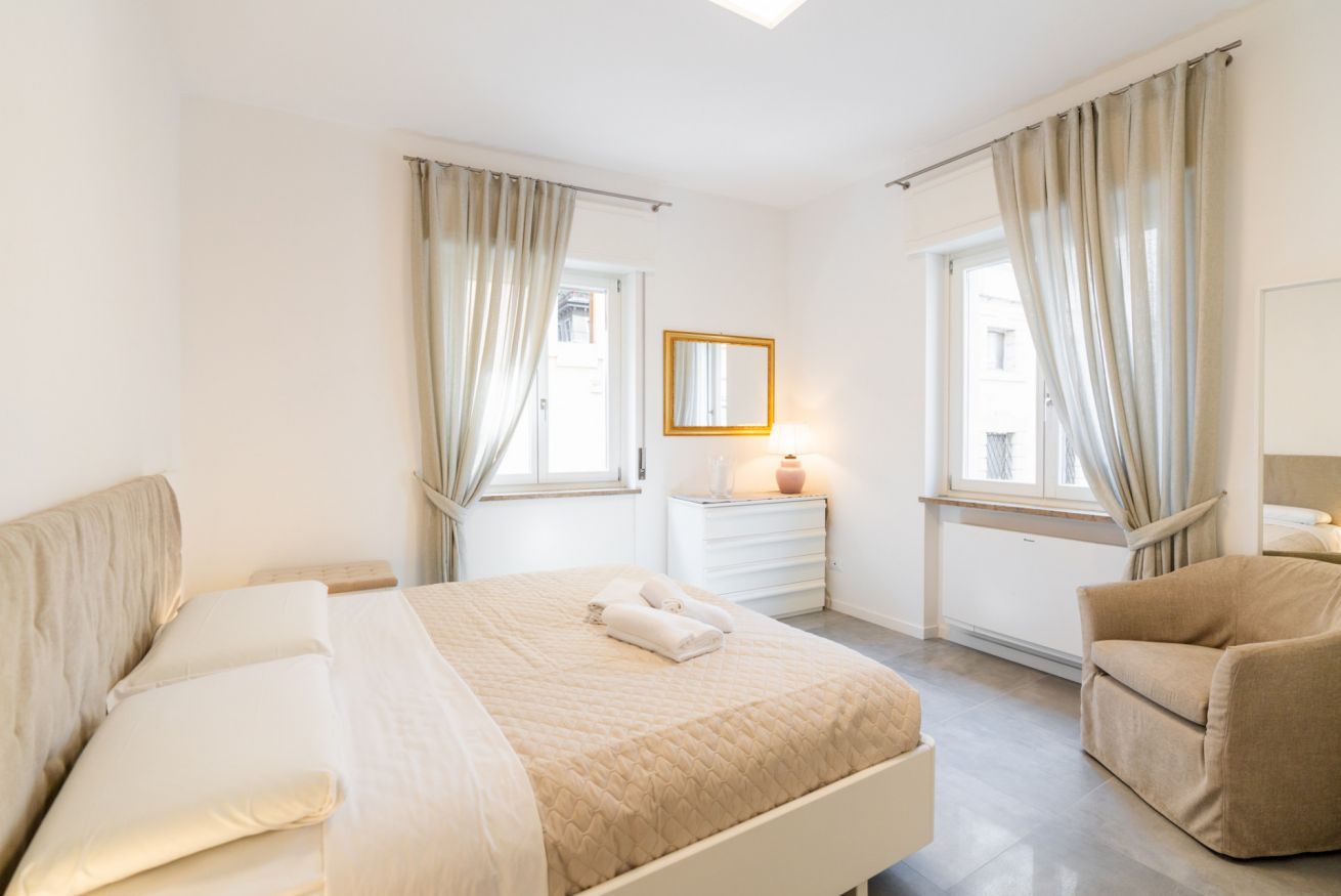 Une chambre à coucher comprenant un lit, une chaise, une table de chevet, un miroir et une fenêtre avec des rideaux.