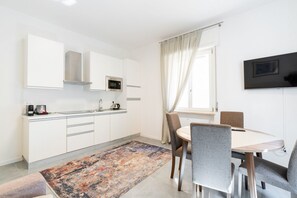 A modern kitchen with white cabinets, a microwave, and a coffee maker. There is a dining table with chairs and a large window with curtains.