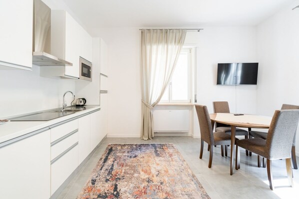 A modern kitchen with white cabinets, a microwave, and a kettle. There's a dining area with a table and chairs, and a TV mounted on the wall.
