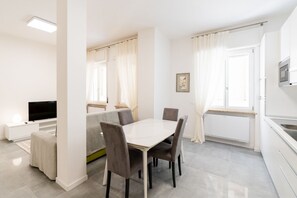 A modern, well-lit room with a dining table, chairs, a sofa, a TV, and a kitchen area. - Melon Court Residence 3 (Verona)