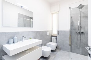 A modern bathroom with a large mirror, a white vanity, a toilet, and a glass-enclosed shower. - Melon Court Residence 3 (Verona)