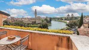 A balcony with a table and chairs overlooking a river with buildings and a tower in the background. - Ponte Pietra Residence (Verona)